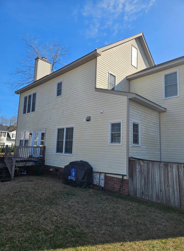 Side view of a two-story beige house with a chimney, deck, and a blue grill cover in a backyard.