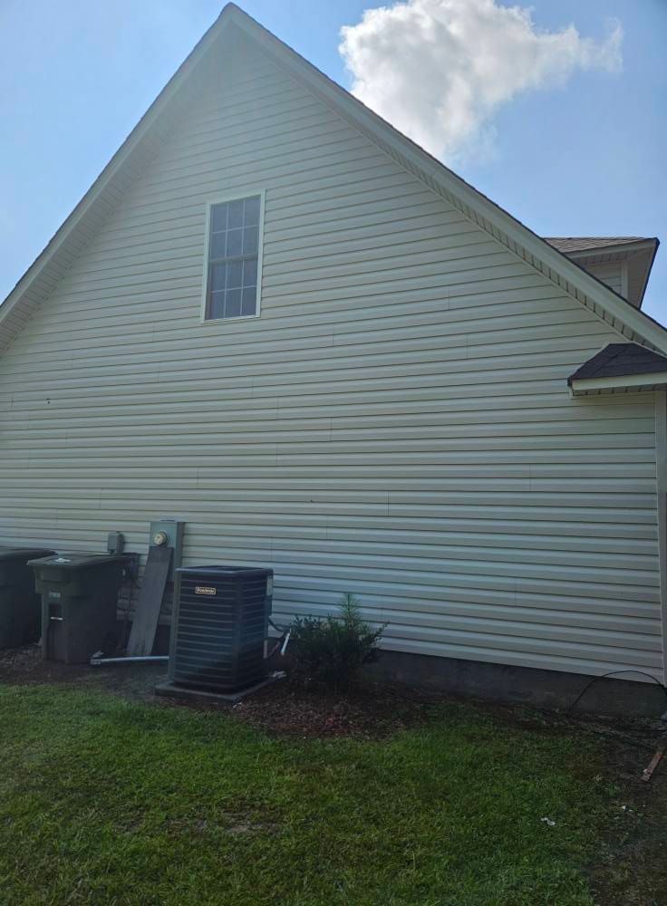 Beige vinyl-sided house with a window, an AC unit, and trash cans. The background is a blue sky and green grass.