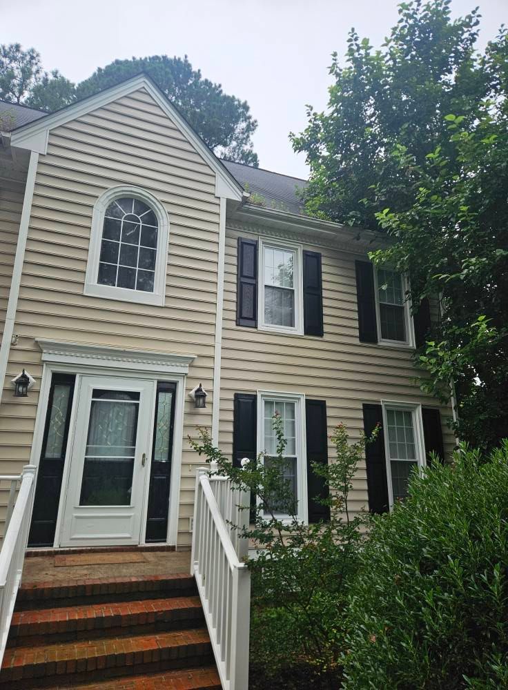 Two-story beige house with black shutters, white trim, front steps, and partial tree coverage.