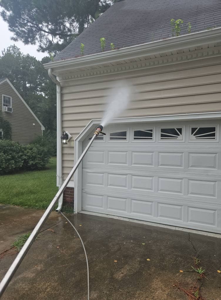 A person power washing a beige house, targeting the siding near the garage.