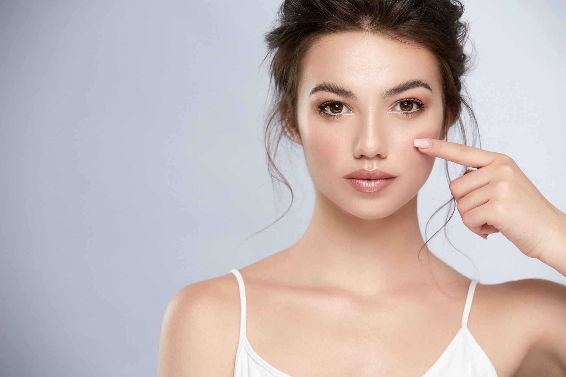 Woman points to her cheek. She wears a white top and has brown hair pulled up. Light background. Woman points to her cheek. She wears a white top and has brown hair pulled up. Light background.