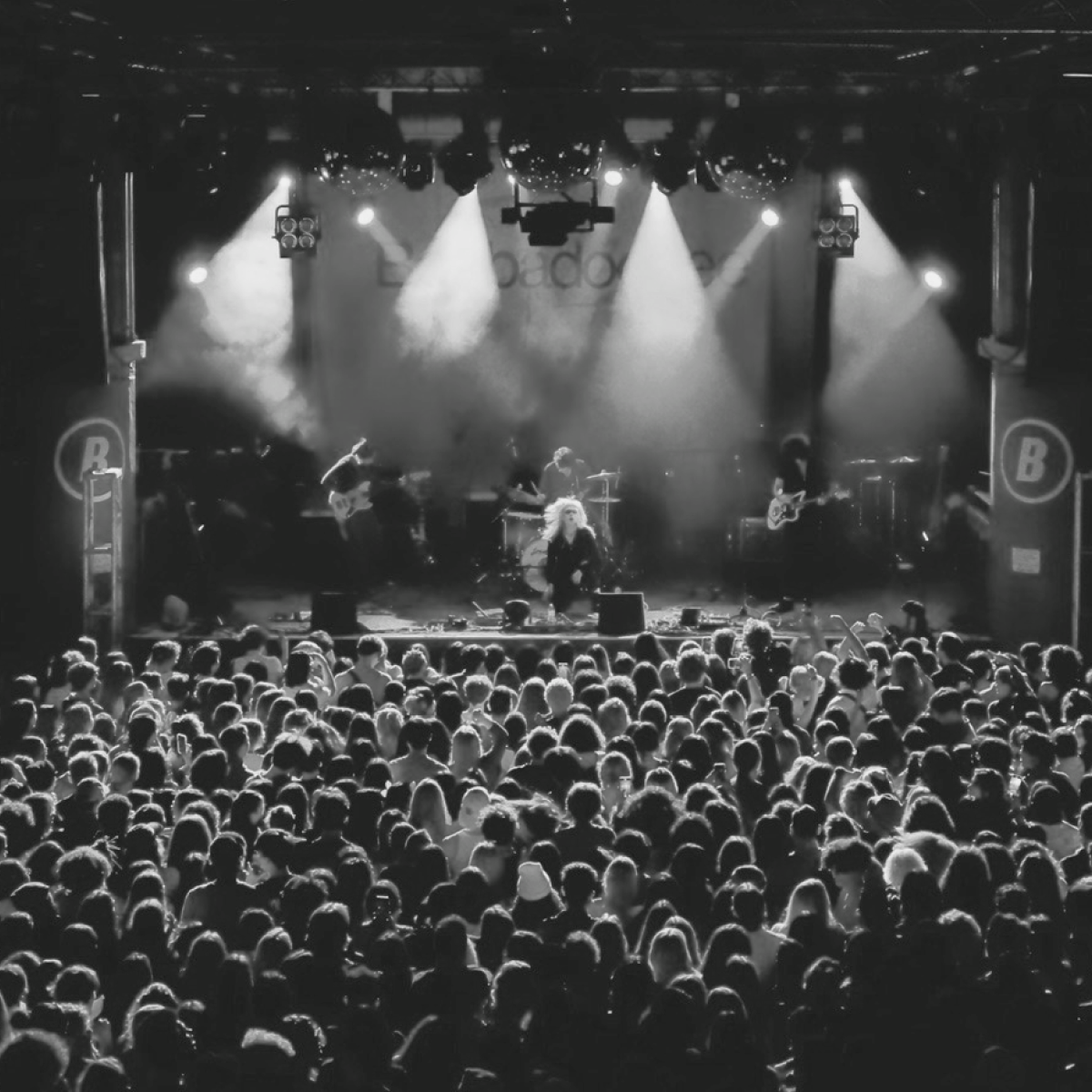 Band performing on stage, bathed in bright lights, to a large, packed audience. Black and white photo.