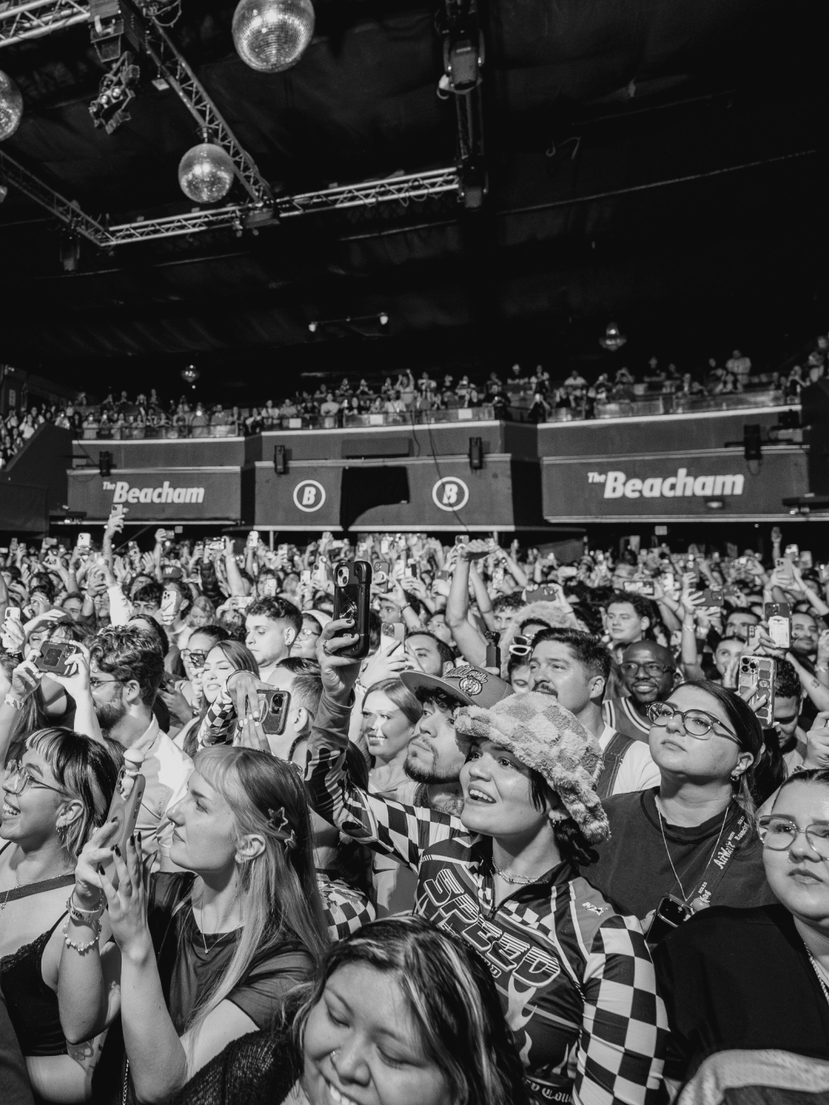 Concert crowd: people in a venue, arms raised, looking up towards stage. Disco balls hang from ceiling.