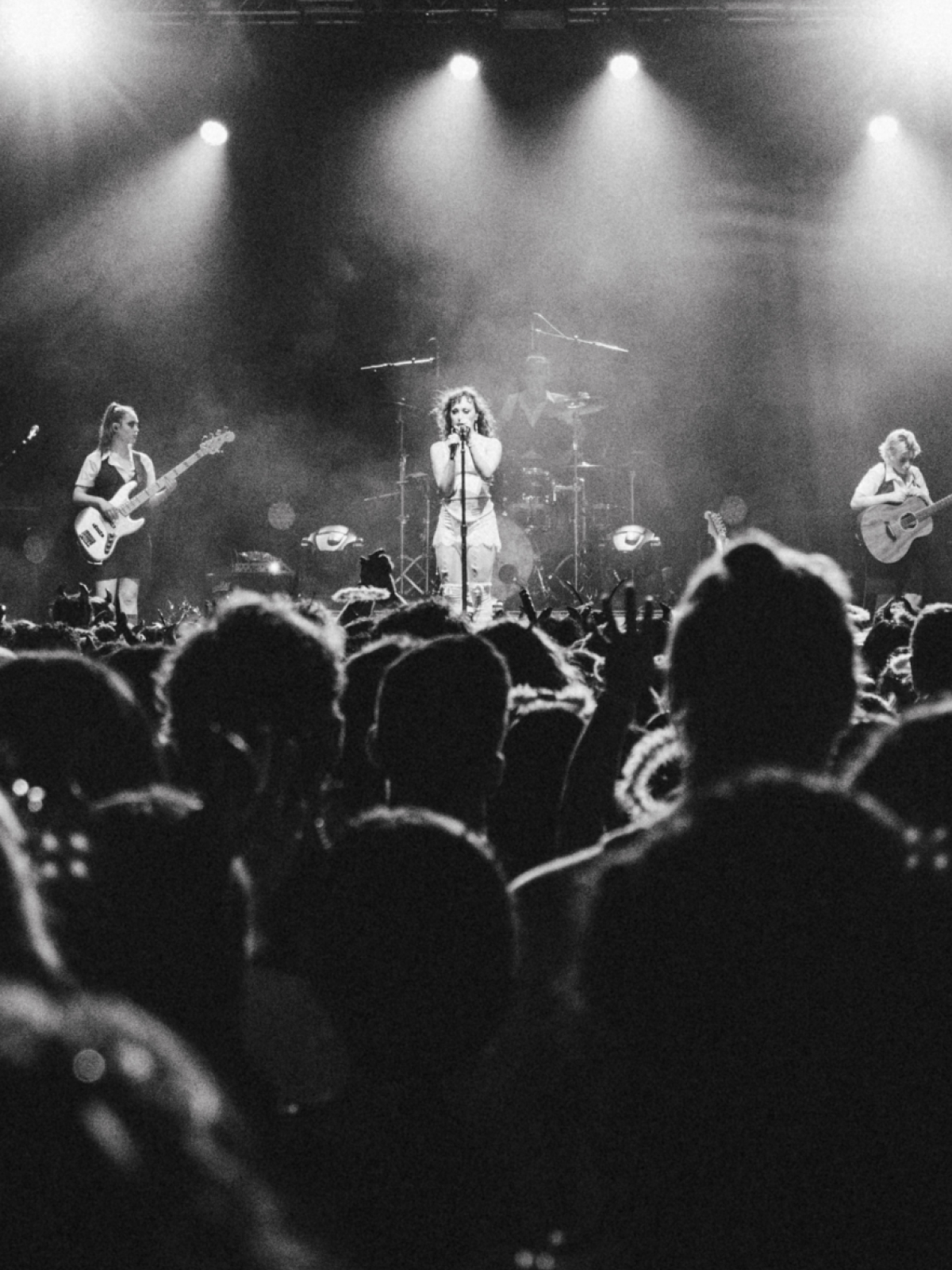 Band performing on stage, bathed in bright lights, with a crowd of people facing them.