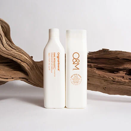 Deux flacons blancs de produits capillaires O&M, l'un étiqueté « Fine Intellect ». Sur un fond de bois flotté.