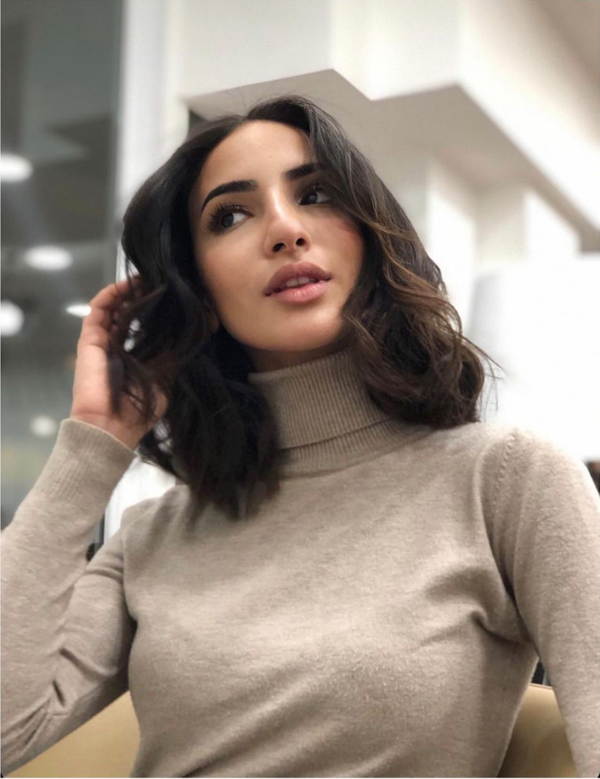 Femme aux cheveux bruns, vêtue d'un col roulé beige, le regard pensif levé, la main dans les cheveux.