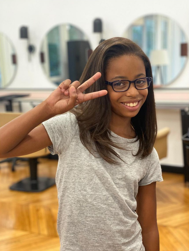 Une jeune fille à lunettes sourit et fait le signe de la paix dans un salon de coiffure avec des miroirs.