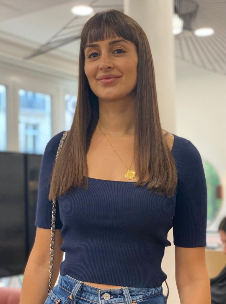 Femme aux cheveux bruns et à frange, portant un haut bleu, souriante, à l'intérieur.