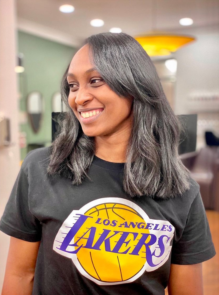 Femme souriante aux cheveux bruns ondulés mi-longs, portant un t-shirt des Lakers dans un salon de coiffure.