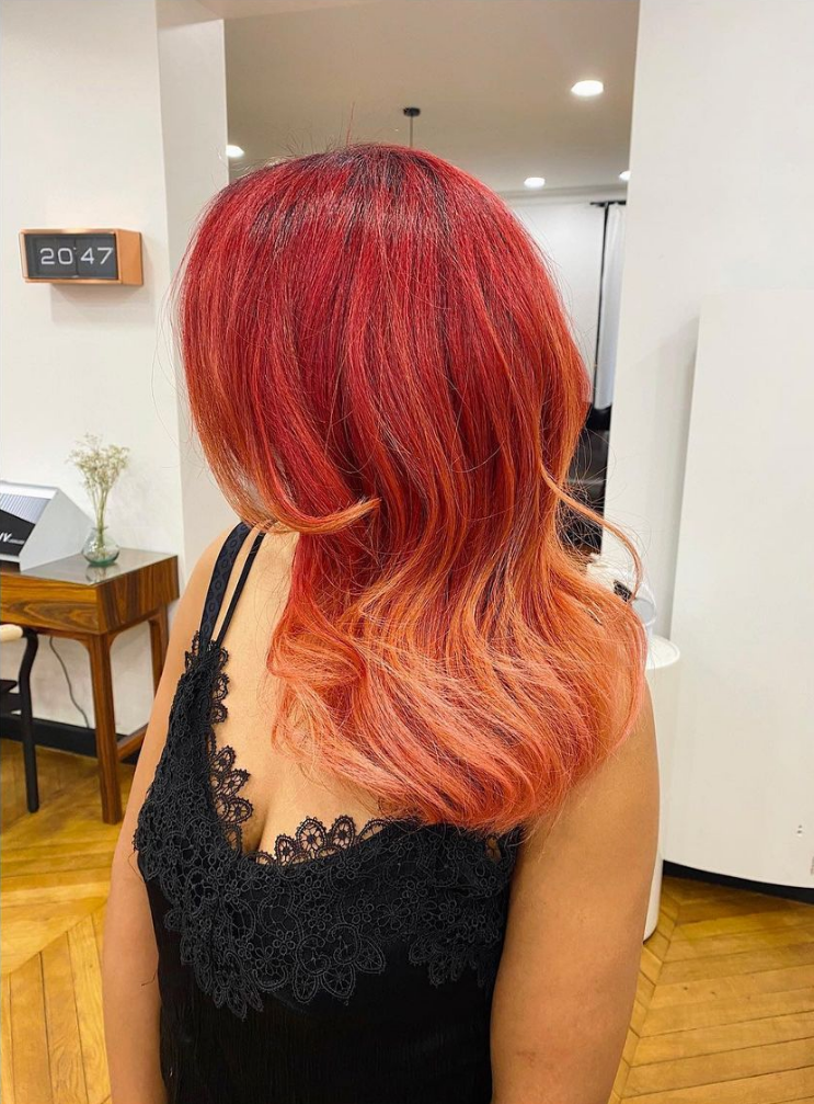 Femme aux cheveux ombrés rouges et pêche dans un salon de coiffure, portant un débardeur en dentelle noire.