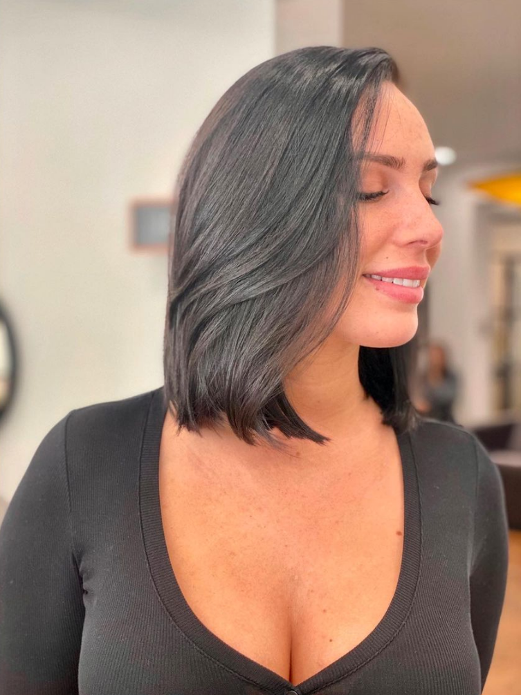 Une femme aux cheveux bruns mi-longs sourit, vêtue d'un haut noir. Dans un salon de coiffure.