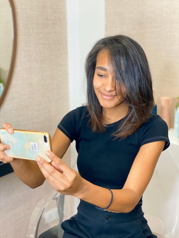 Femme en chemise noire prenant un selfie avec son smartphone, souriante. Ambiance salon avec miroir et chaise.
