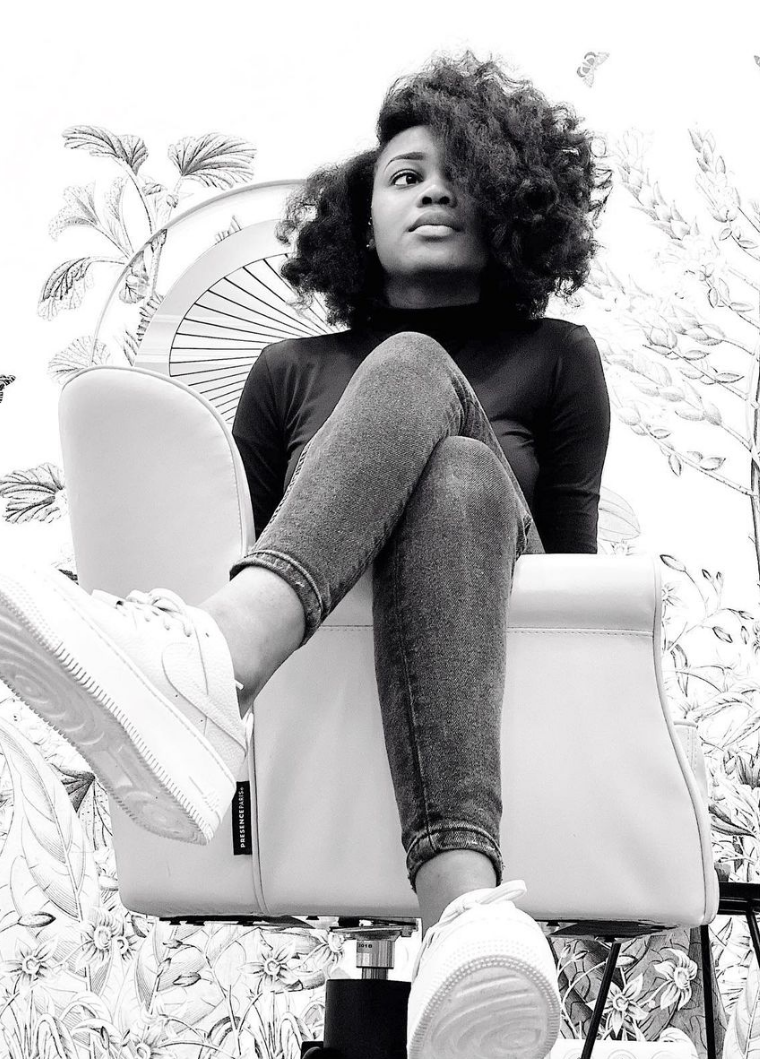 Femme assise, jambes croisées, vêtue d'un col roulé noir, d'un jean et de baskets. Elle se détache sur un mur à motifs.