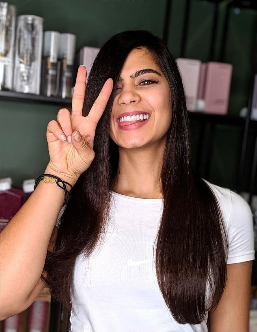 Une femme aux cheveux foncés sourit en faisant le signe de la paix. Chemise blanche, étagères en arrière-plan.