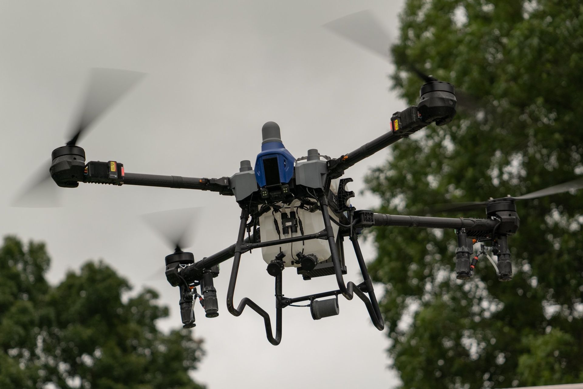 Drone in flight, spraying liquid over a field. Gray body, black arms, white tank.