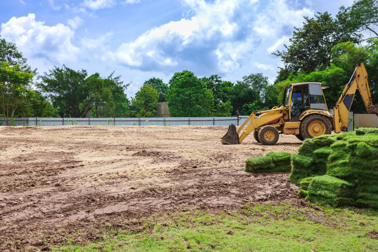 Yellow construction vehicle on a cleared lot with muddy ground, stacks of sod, and trees under a cloudy sky.