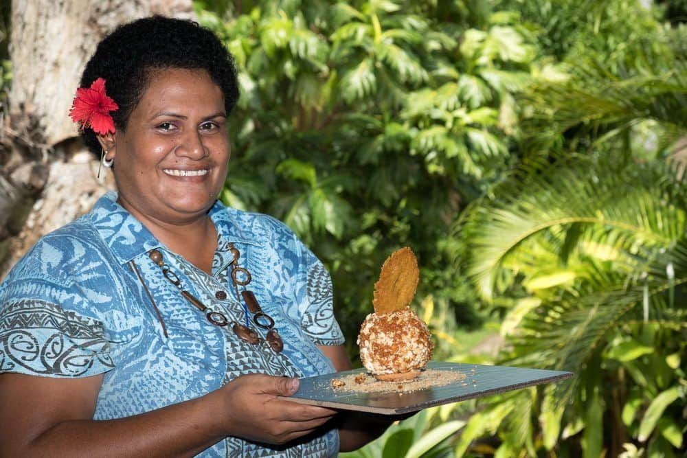 A Fijian lady delivering a drink on a tray in front of sunshine lit tropical plants 