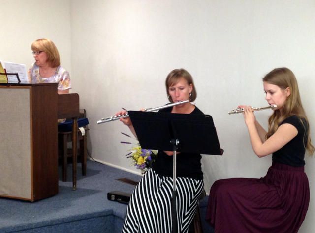 Two flutists perform beside a podium while a woman speaks at a lectern on a small stage.