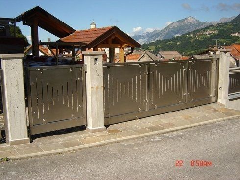 Un cancello metallico con aperture verticali si erge tra due pilastri di pietra, con un villaggio di montagna sullo sfondo.