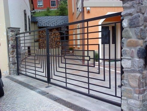 Un grande cancello carrabile in metallo nero con un design geometrico a telai rettangolari incastrati, fiancheggiato da pilastri in pietra.