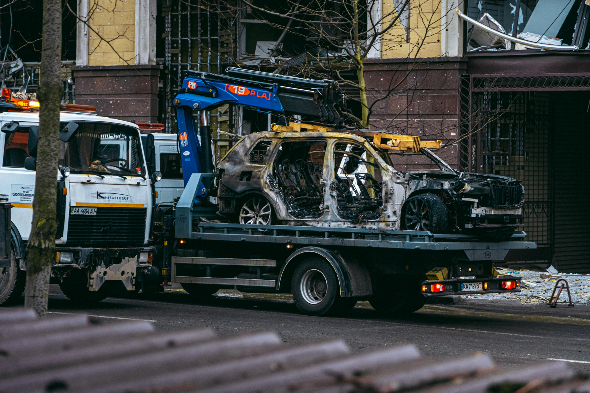 A burned-out car on a tow truck. Damaged building in the background.