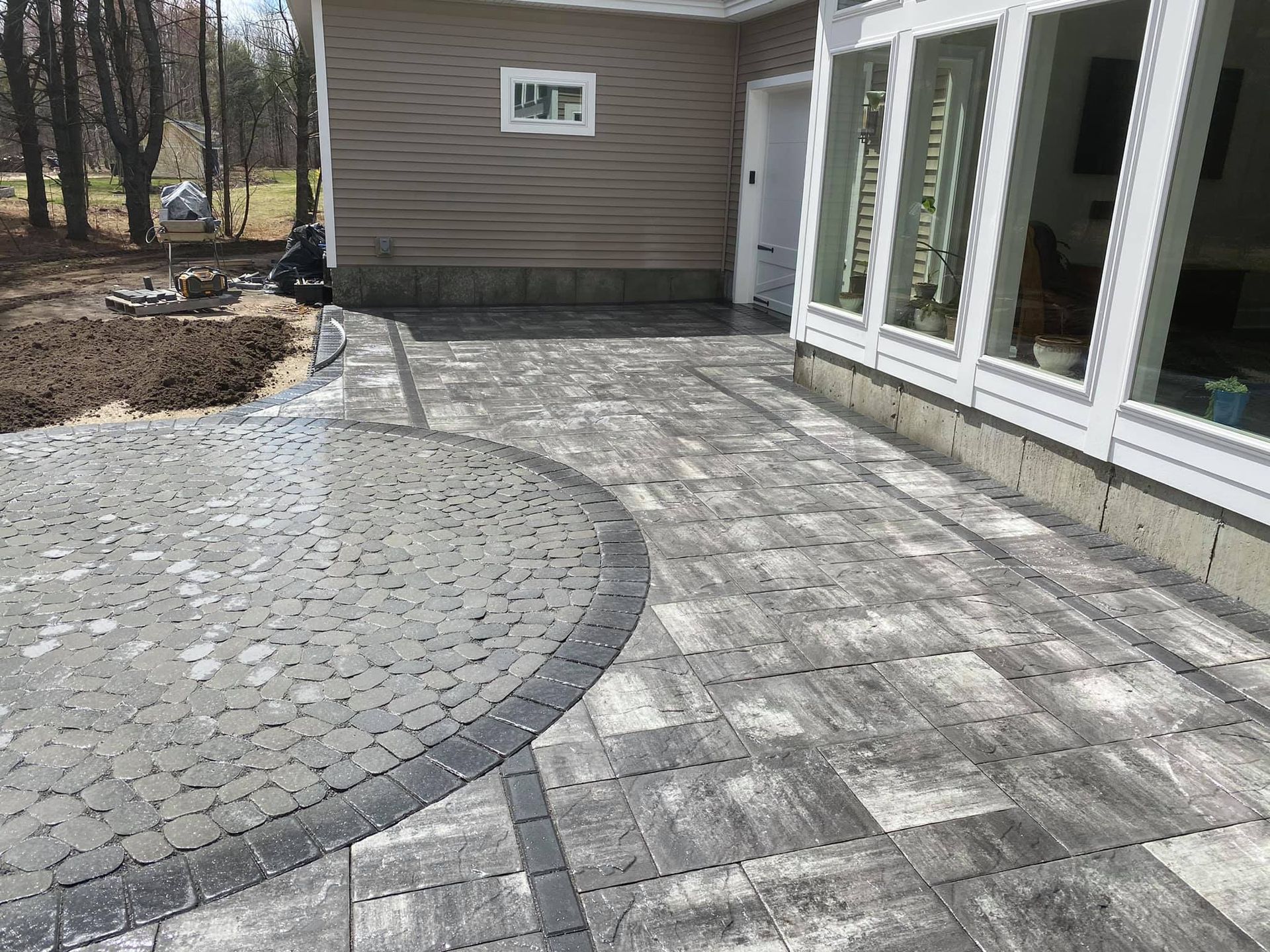 Stone patio with curved edge, bordered by dark bricks, next to a house with large windows.