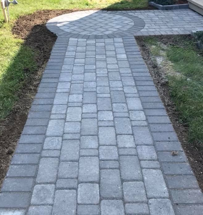 Brick pathway with a circular landing, bordered by grass. Gray brick pavers in a formal garden.