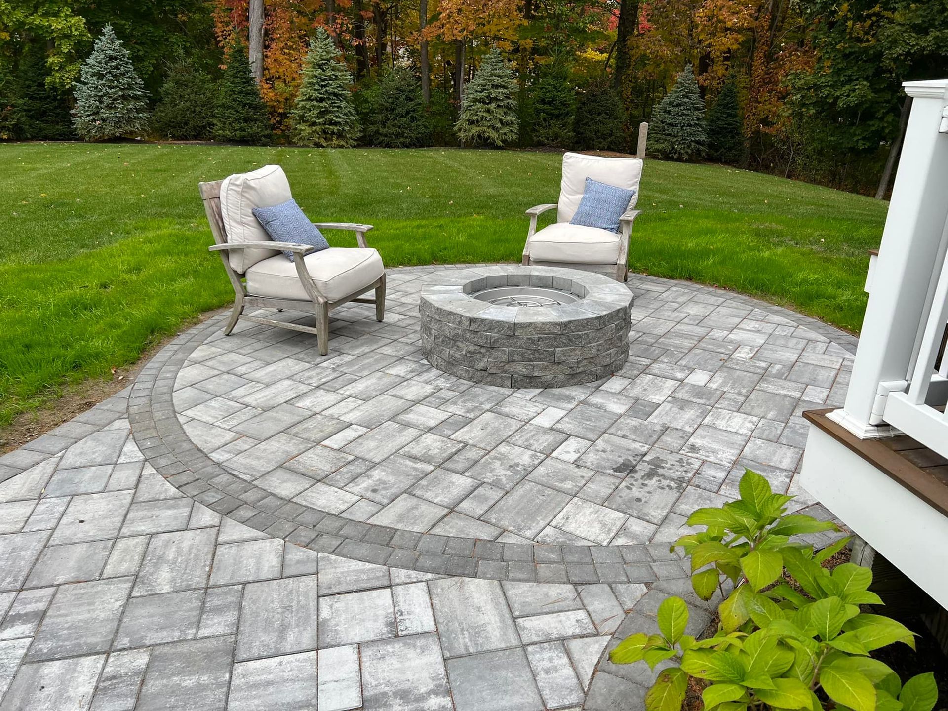 A circular patio with a fire pit and two chairs, surrounded by grass.