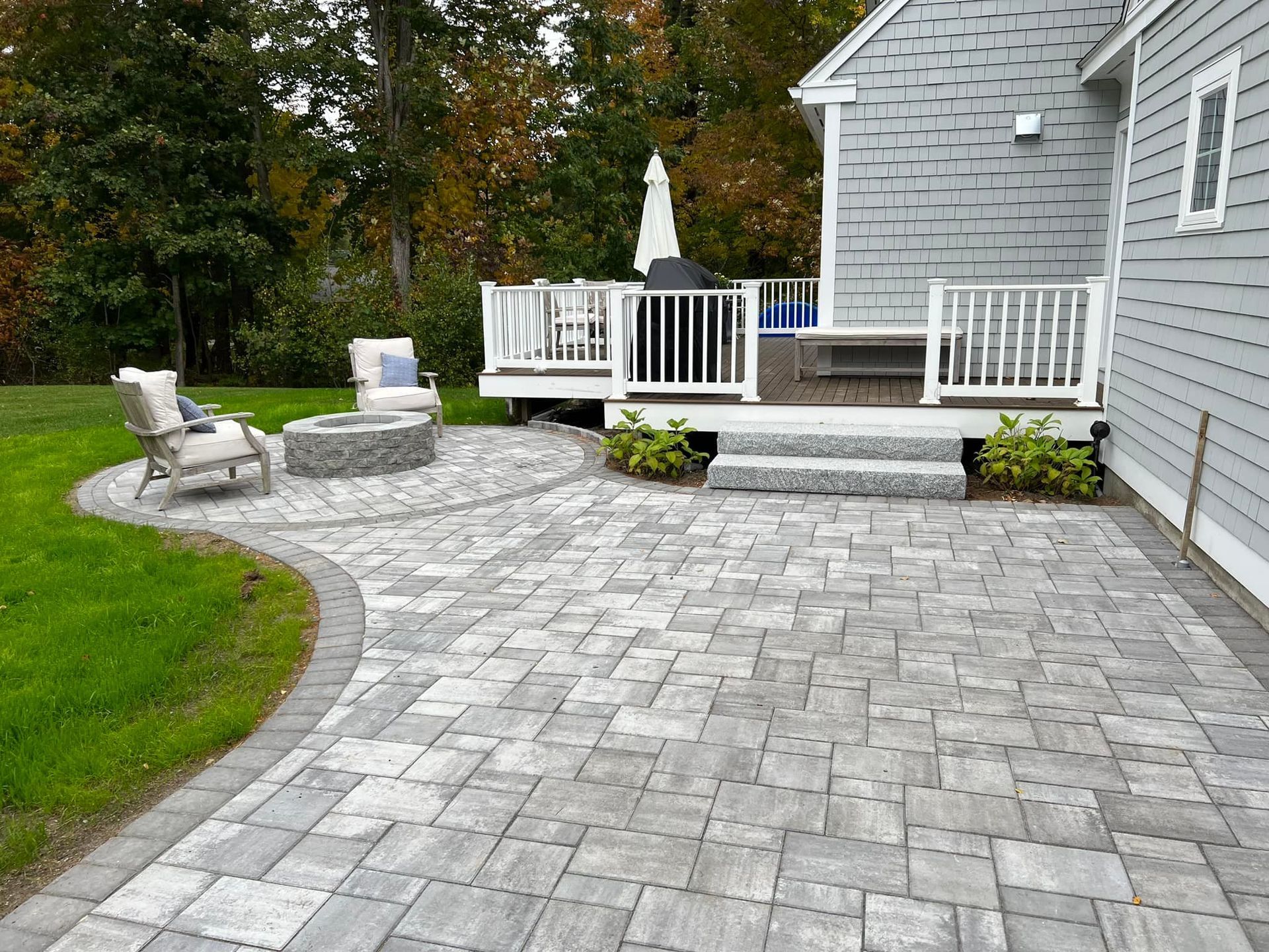 A brick patio with a fire pit and deck. Gray pavers and a gray house. Two chairs.