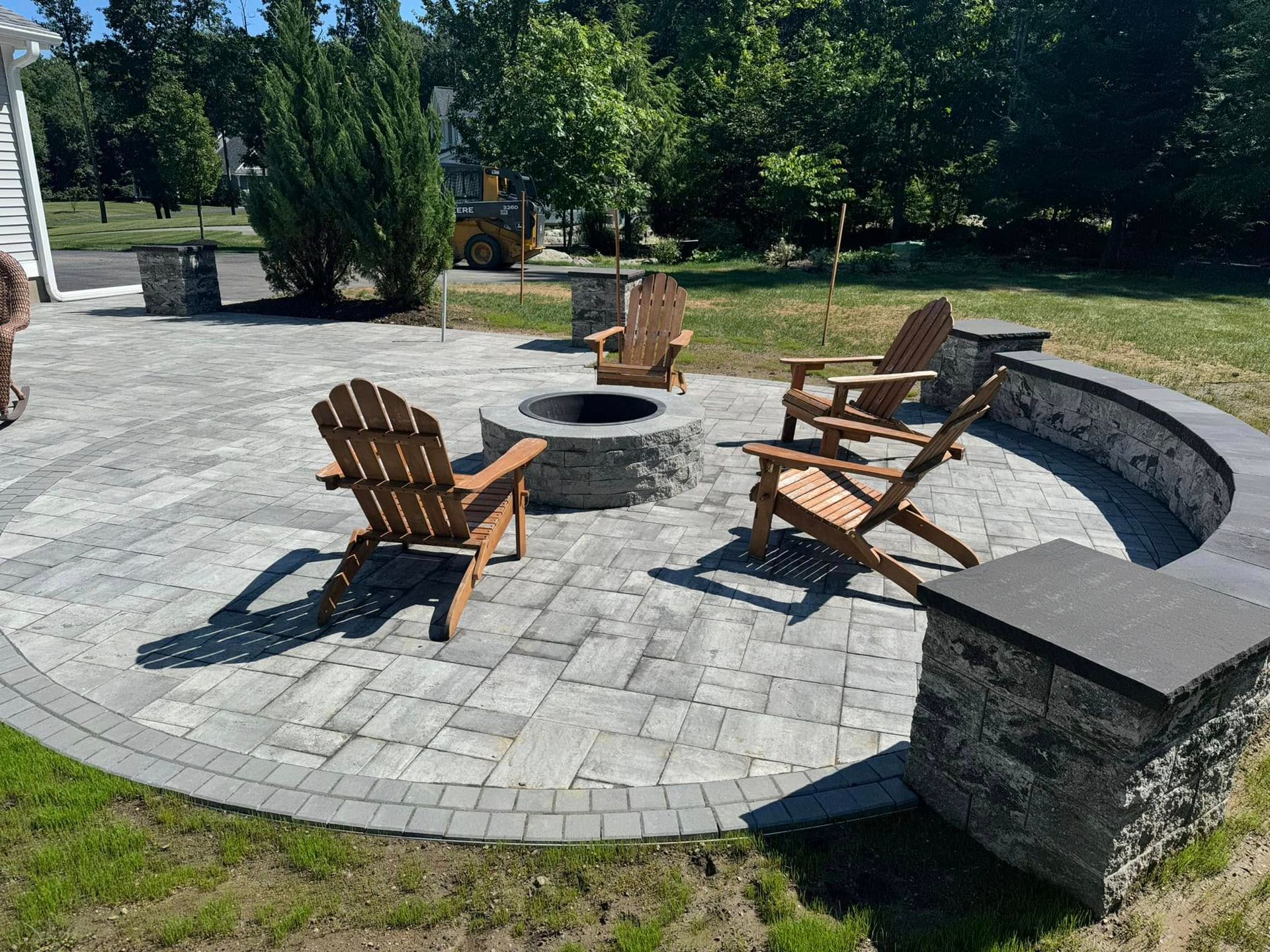 A brick patio with Adirondack chairs around a fire pit. Stone walls, trees, and green grass surround it.