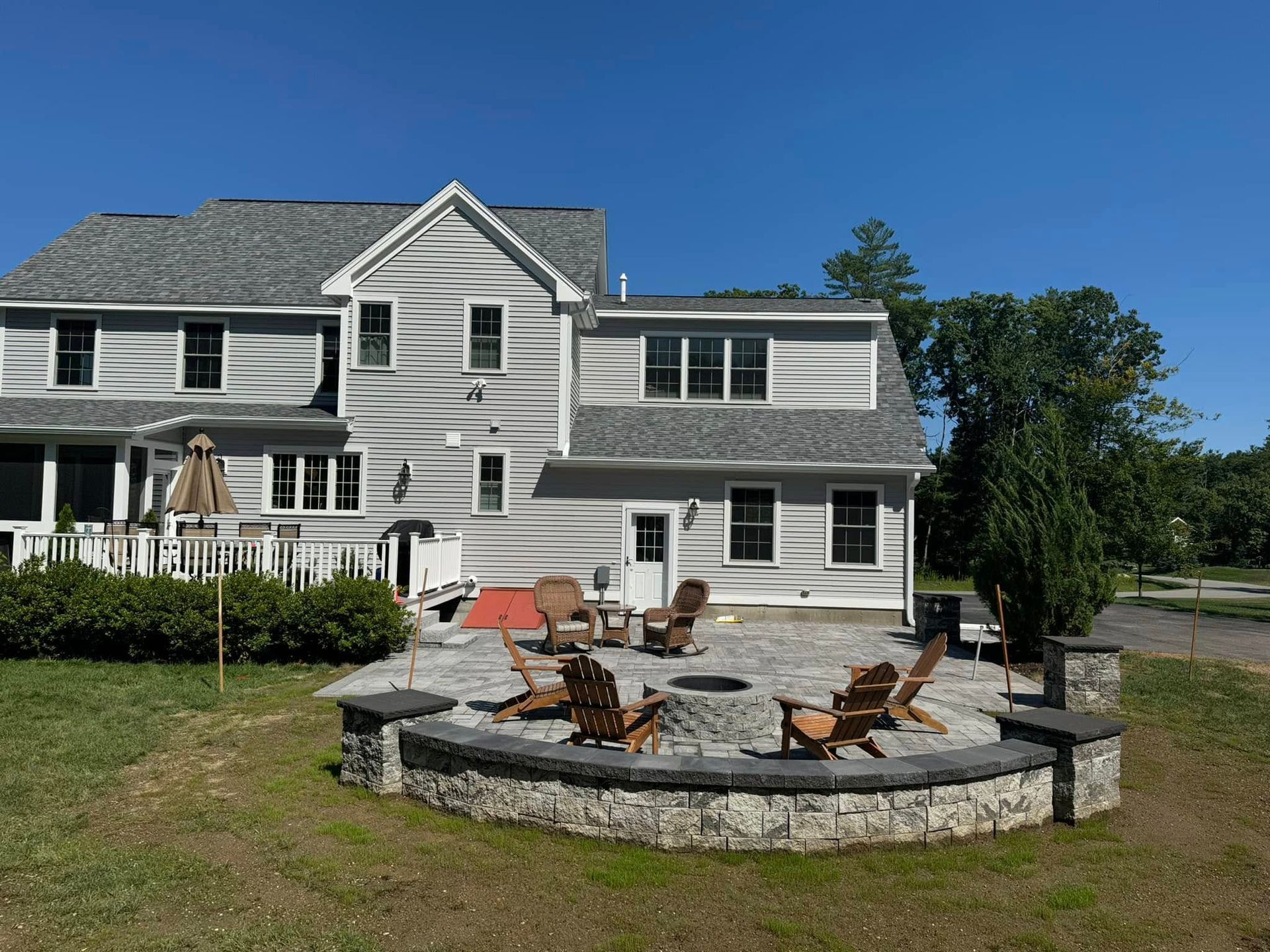 Backyard with stone fire pit and wooden chairs, next to a gray house under a clear blue sky.