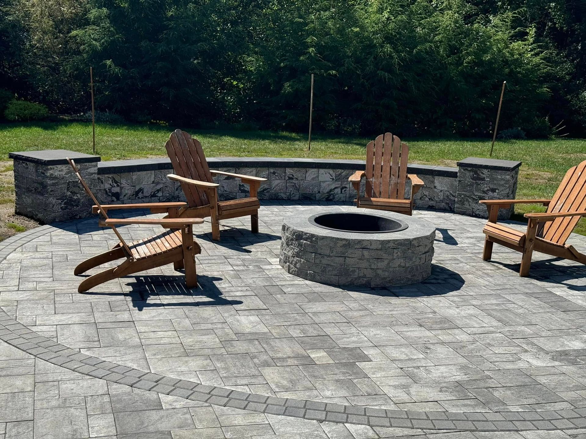 Patio with Adirondack chairs around a stone fire pit, framed by low stone walls, green trees in the background.