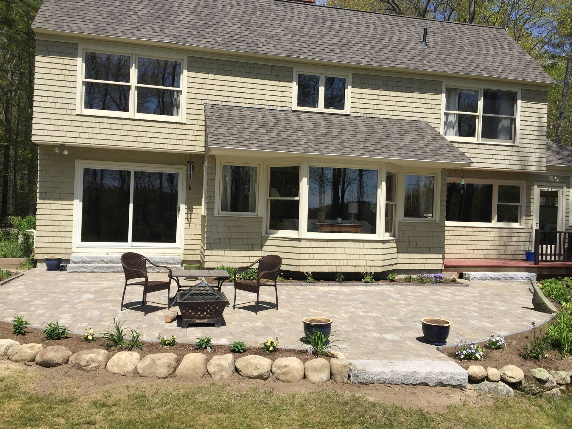Two-story beige house with a paved patio, fire pit, two chairs, and rock garden in a sunny outdoor setting.