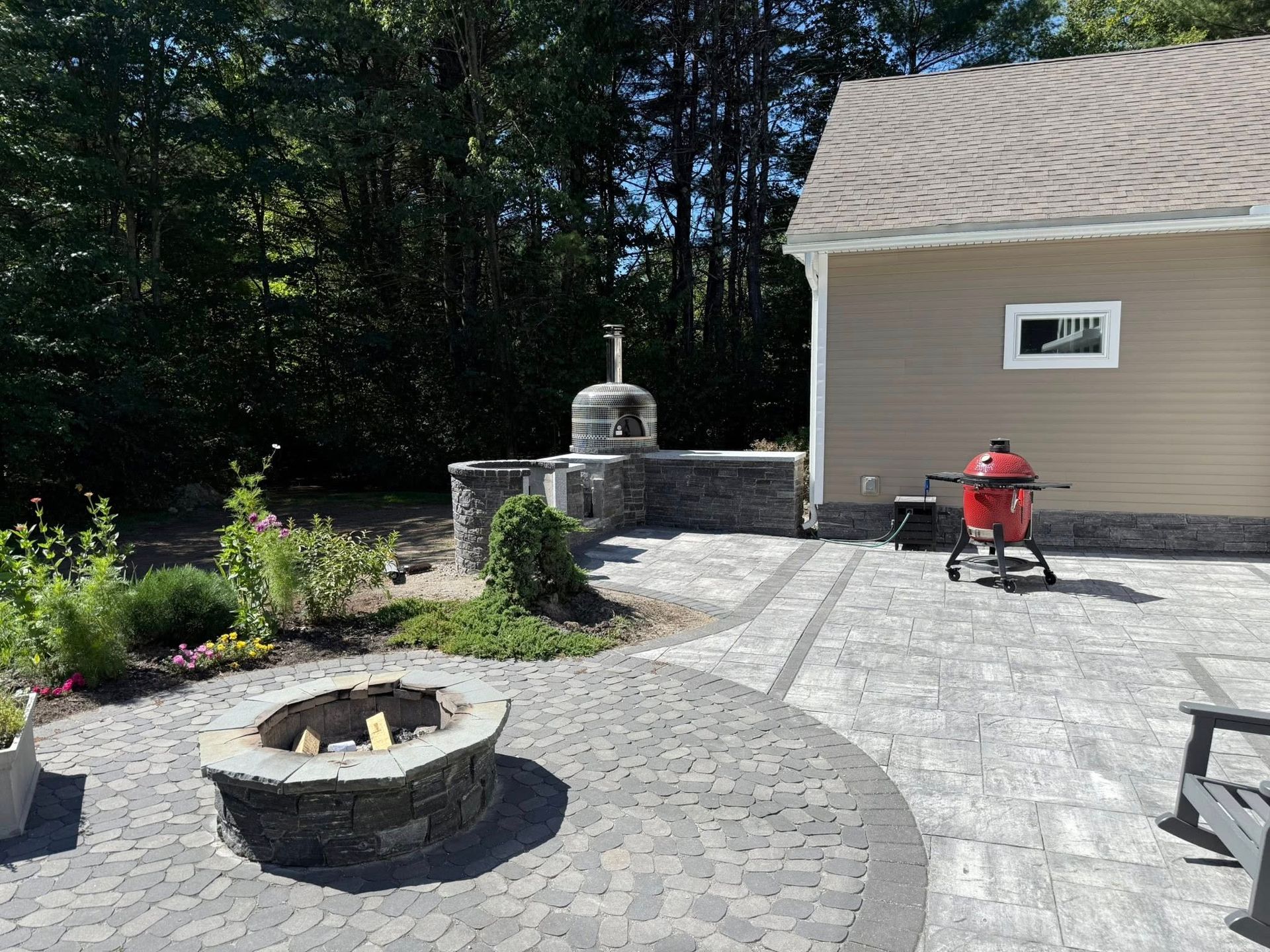 Backyard patio with fire pit, pizza oven, grill, and house; sunny day.