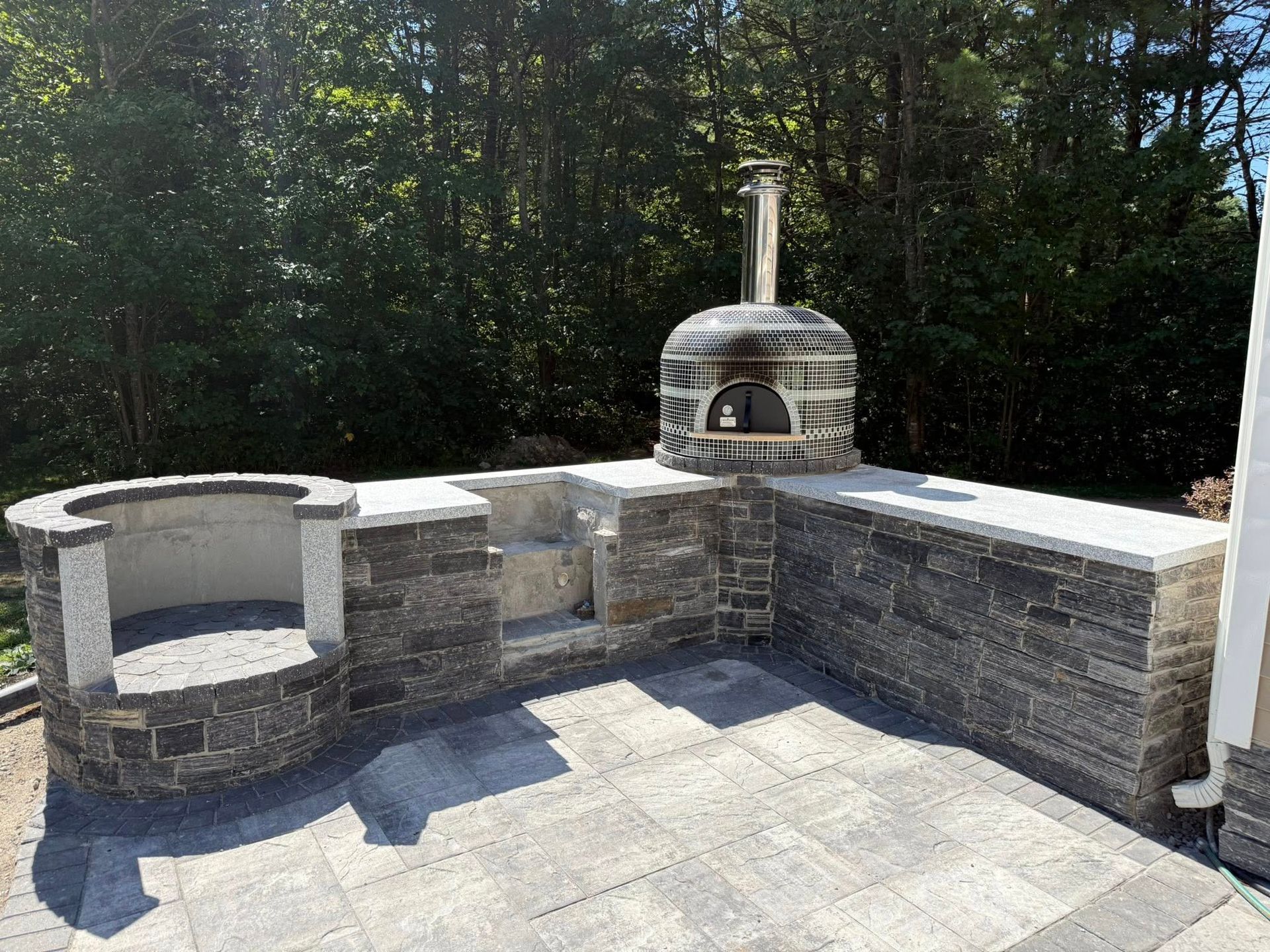 Outdoor kitchen with stone oven, grill, and counters on a paved patio, backed by trees.