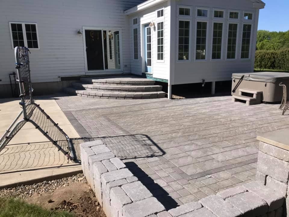 Backyard patio with paving stones, retaining wall, and hot tub. House with multiple windows and steps leading to a sunroom.