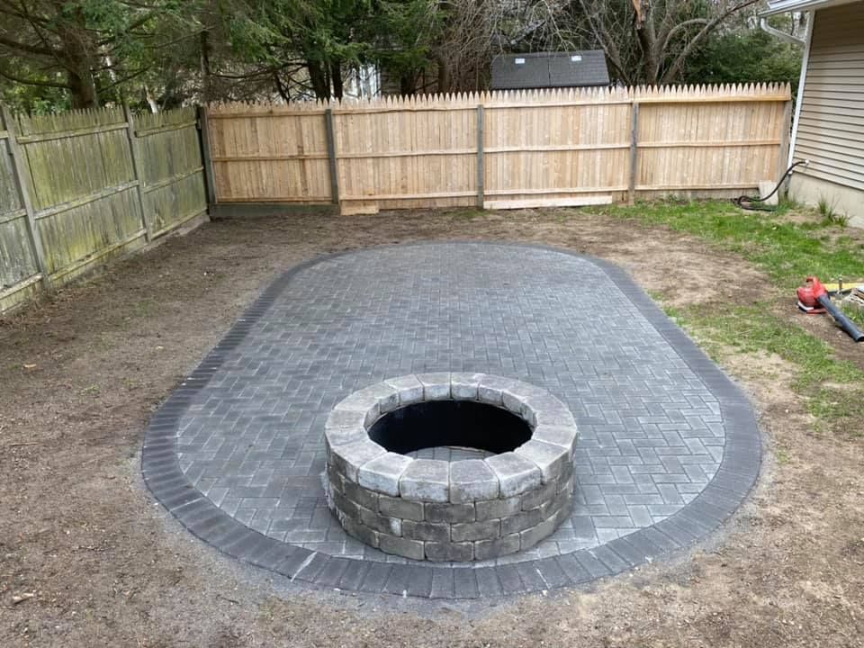 A gray stone fire pit on a paved patio in a backyard, bordered by a light brown fence.