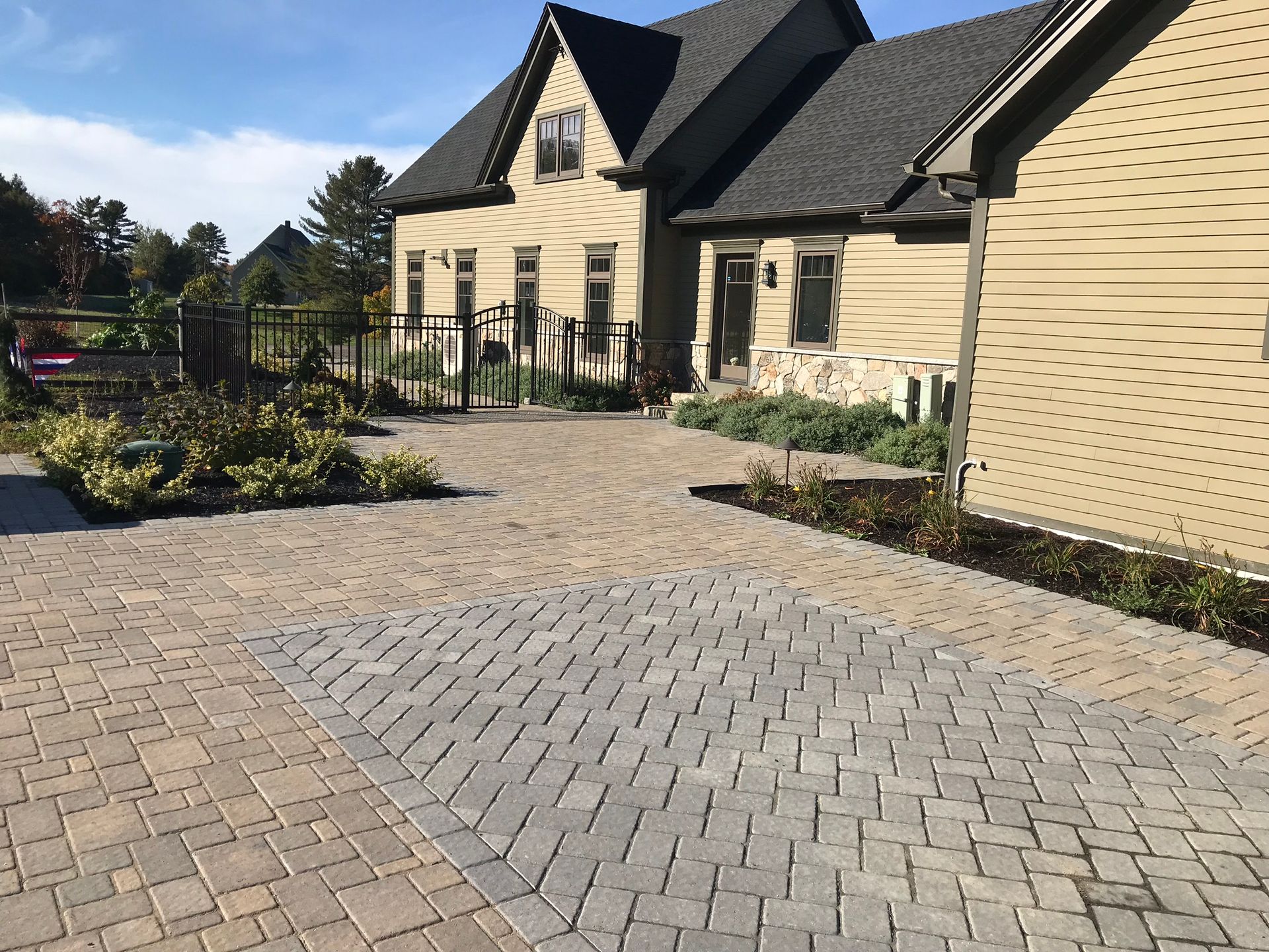 Brick paved driveway leading to a tan house with a dark roof, surrounded by landscaping.