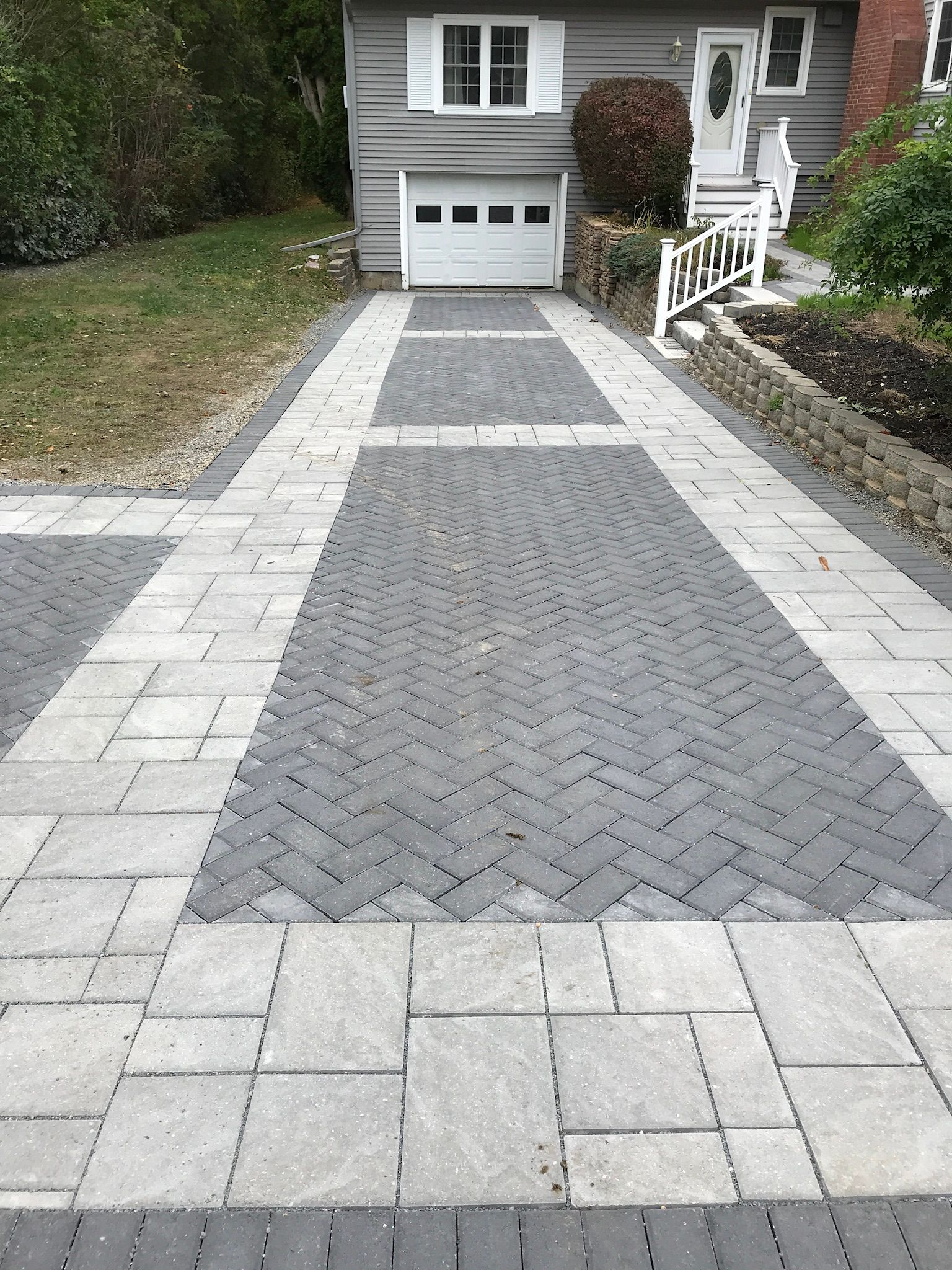 Brick paver driveway leading to a gray house with a white garage door and front door.