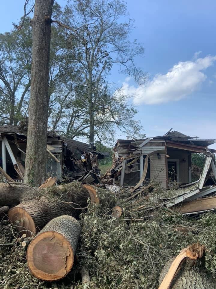 Damaged houses amid fallen trees and debris under a partly cloudy sky.