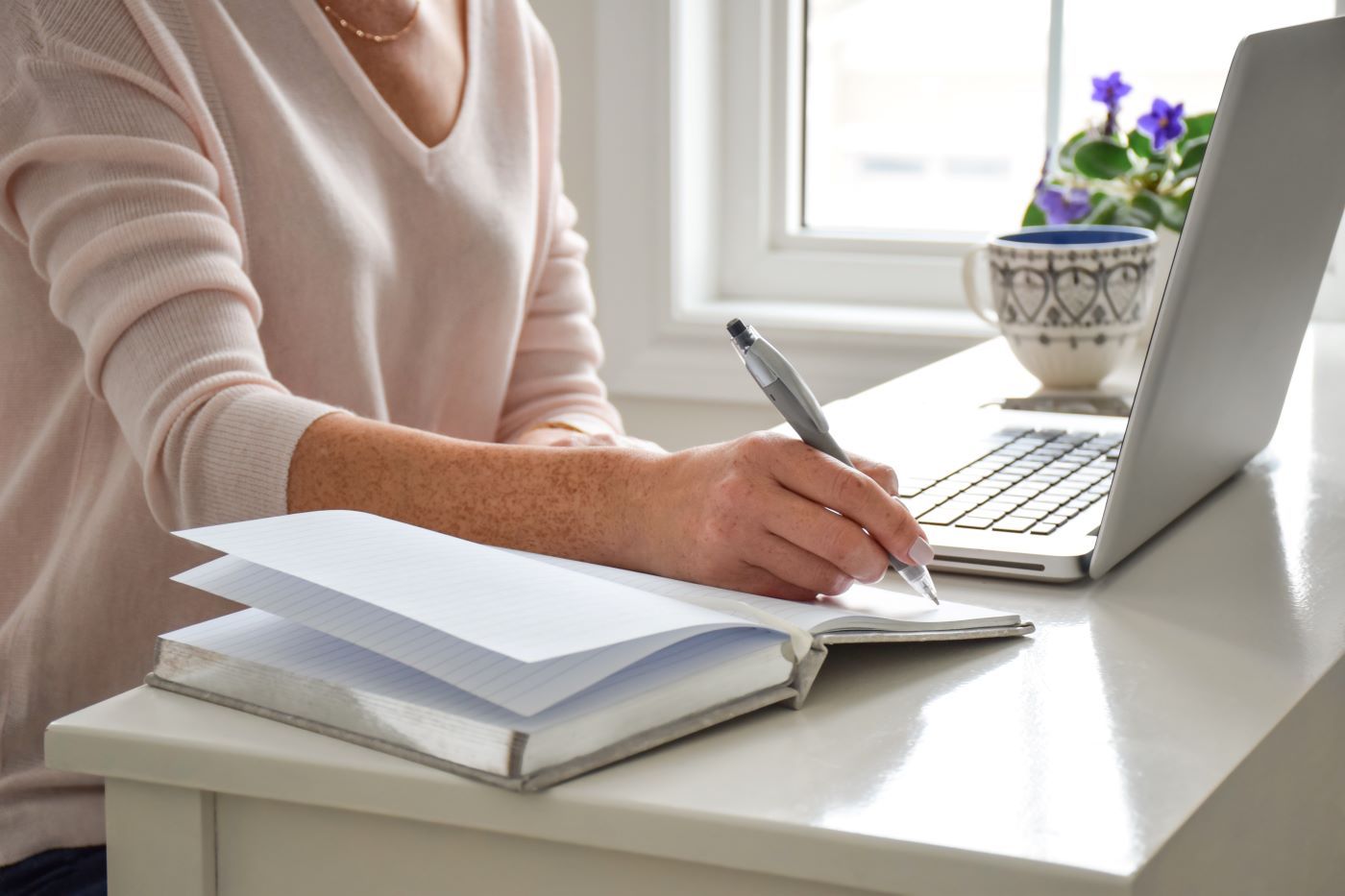 A woman is writing in a notebook in front of a laptop computer.