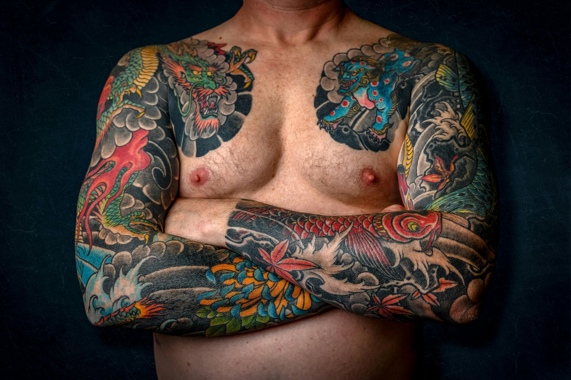 Torso tatuato con le braccia incrociate, che mostra colorati tatuaggi su maniche e petto su uno sfondo scuro.