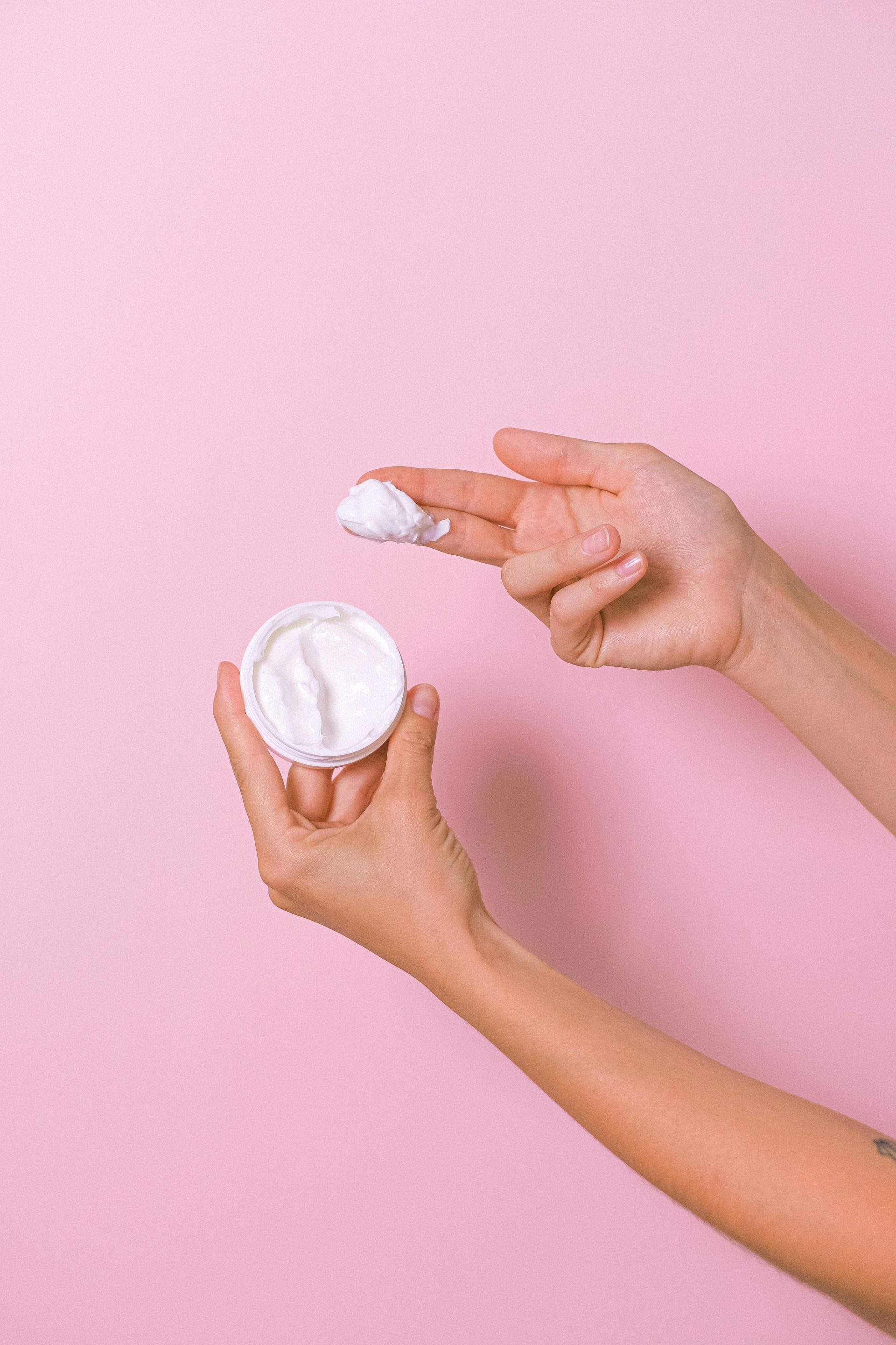 Mani che applicano crema bianca da un vasetto su uno sfondo rosa.