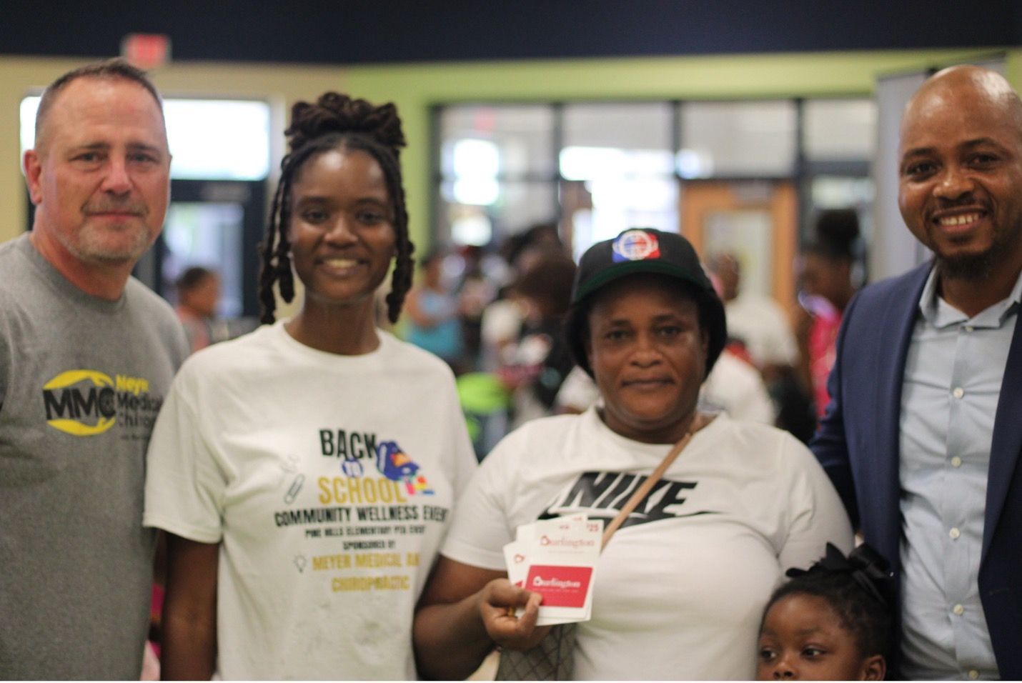Group photo at a back-to-school event. People smiling, holding items. Indoor setting, light background.