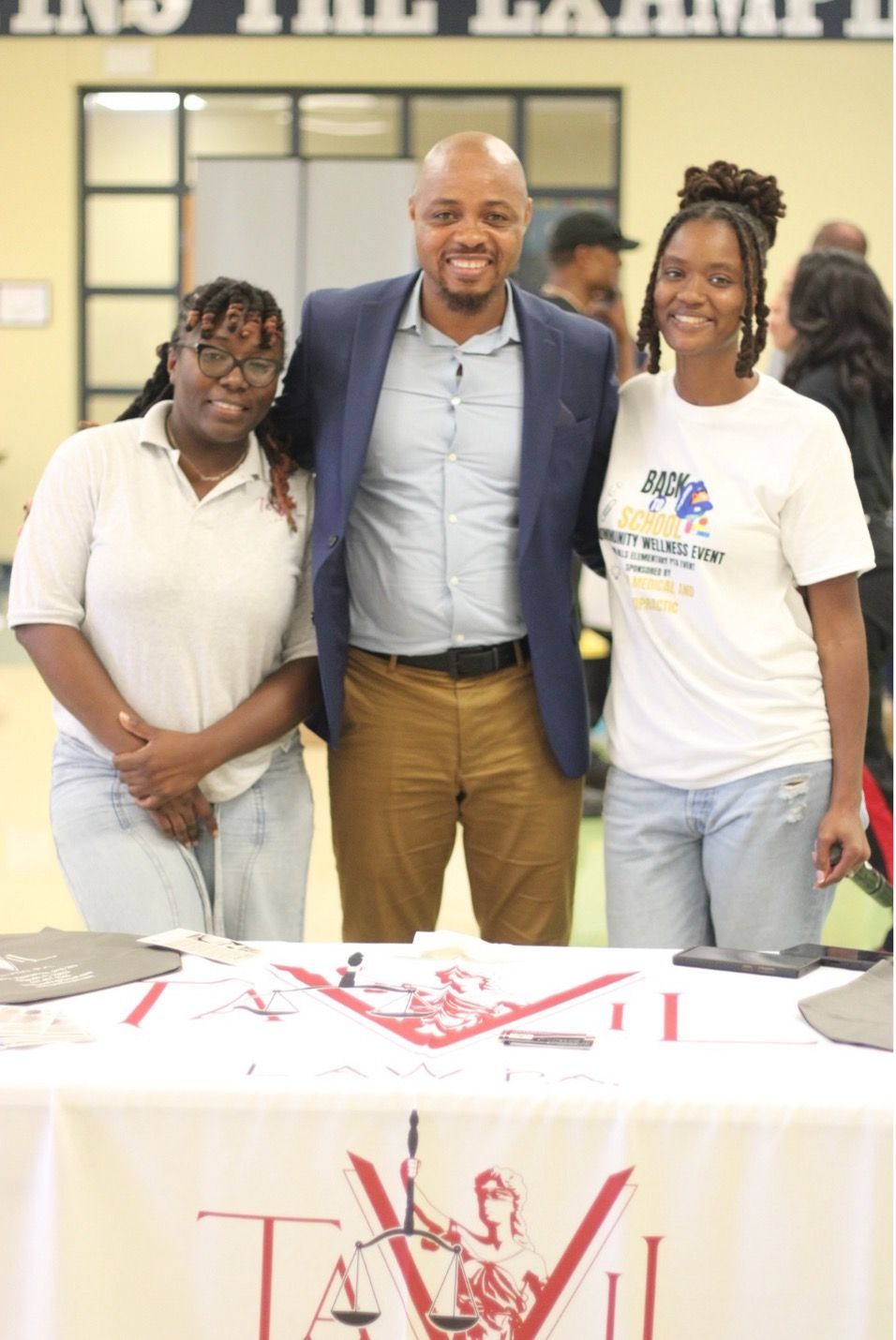 Three people pose together behind a table with a logo; man in blazer, two women in t-shirts; indoor setting.