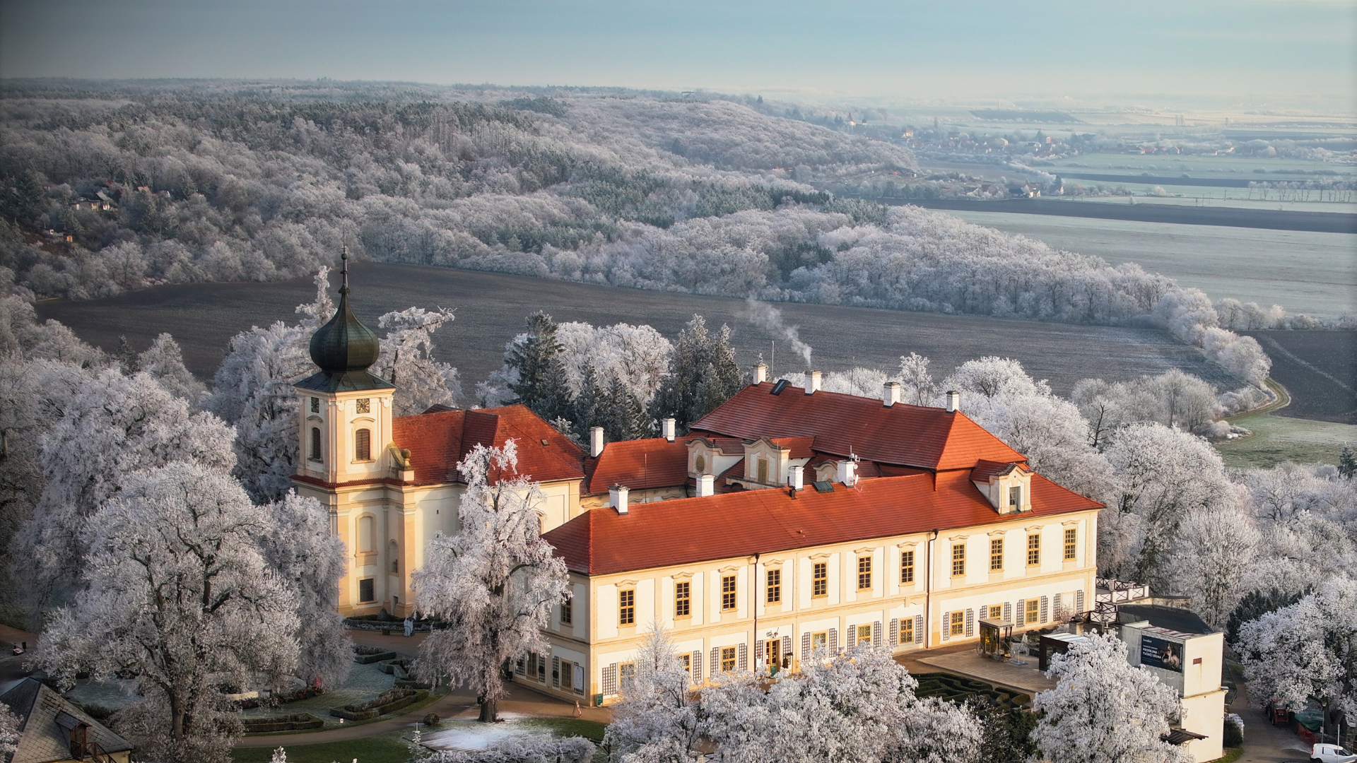 Velká bílá budova se zvonicí je obklopena stromy a trávou.