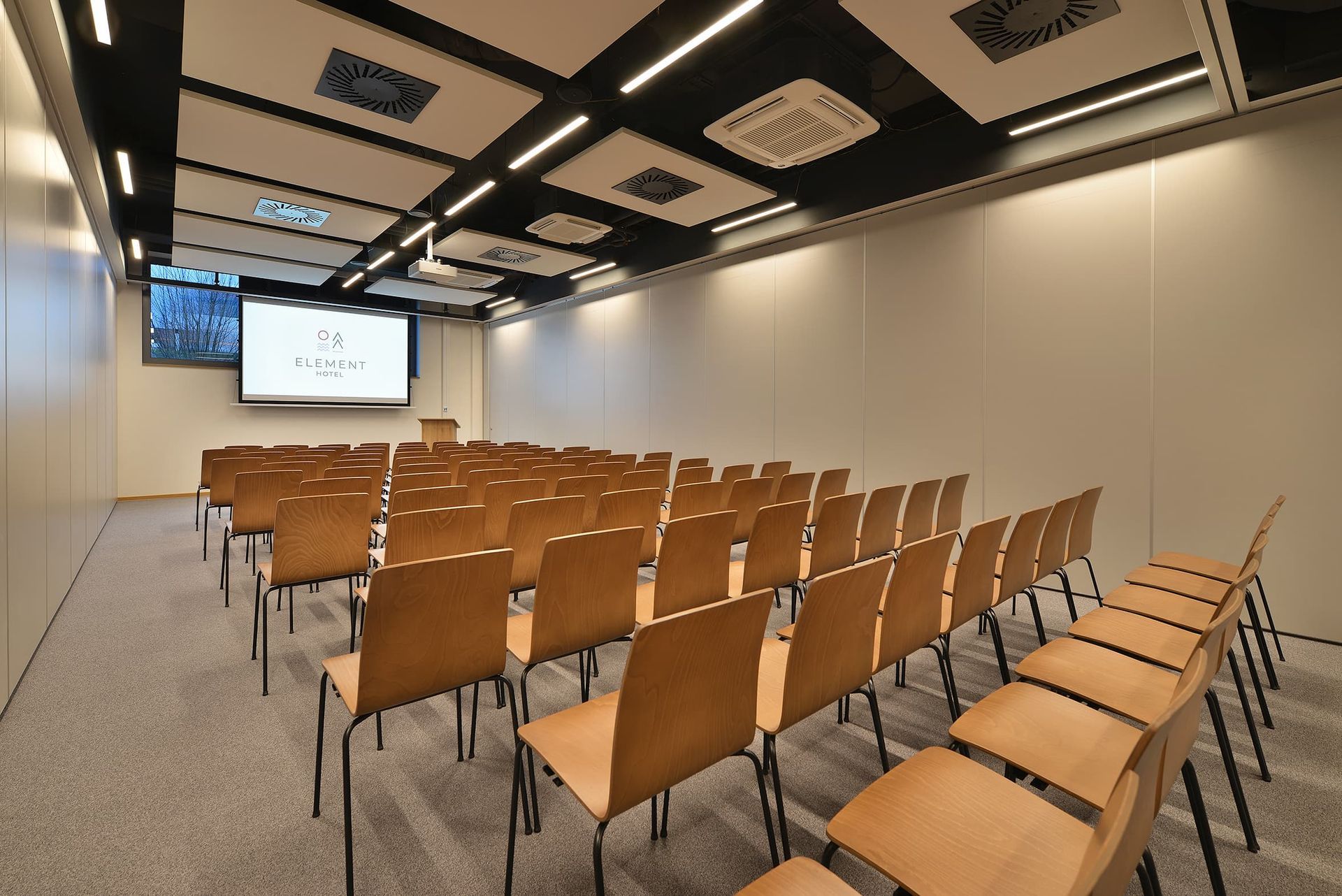Konferenční místnost s řadami dřevěných židlí a velkou obrazovkou