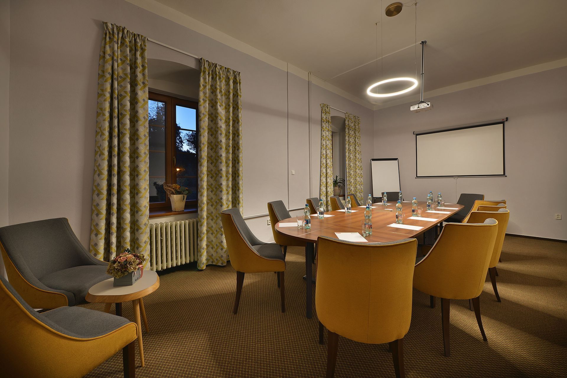 Konferenční místnost s dlouhým stolem a židlemi a promítacím plátnem.