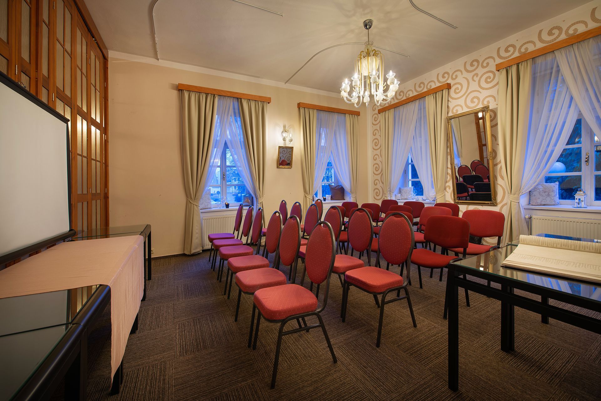 Konferenční místnost s červenými židlemi, stoly a promítacím plátnem.