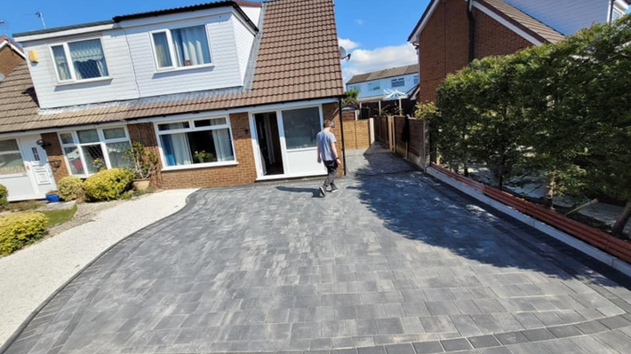 A person walks toward the front entrance of a two-story house with a newly laid grey block-paved driveway.